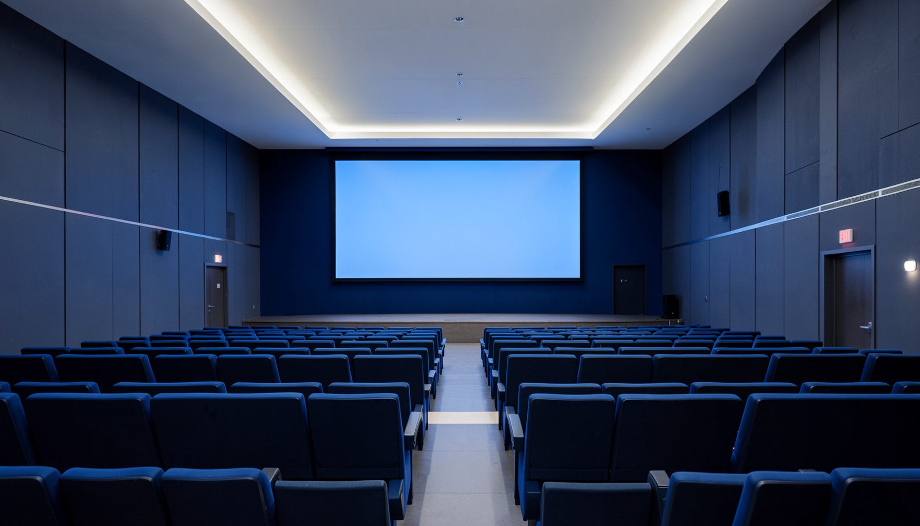 Auditorio moderno con butacas en tonos azul oscuro, iluminación cenital suave y una gran pantalla de presentación al fondo, atmósfera académica y profesional.